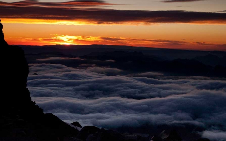High above clouds, the sun rises or sets, causing the sky to appear in shades of orange and yellow.